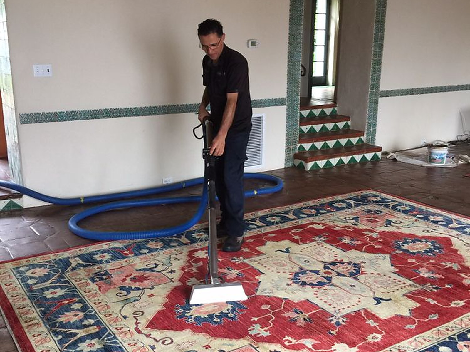 A man drawing a cleaning wand across a red and blue oriental rug, the hose running out through the doorway