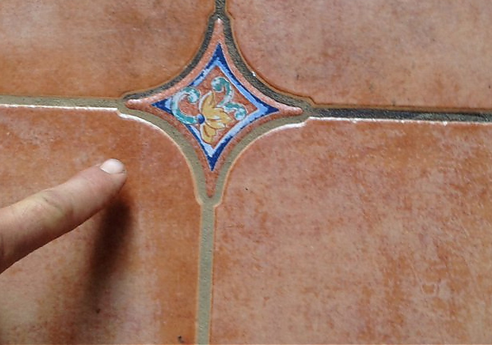 A hand at the grout line where four terracotta tiles meet: pale clean grout to the left of a painted insert, dark soiled grout to the right